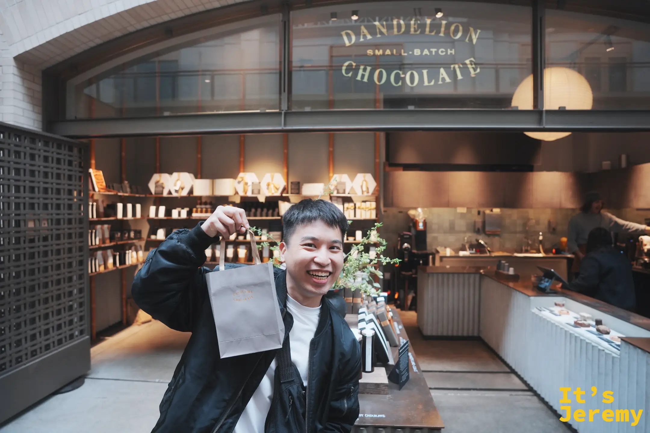 Jeremy 站在 Dandelion Chocolate 巧克力店前微笑，手裡提著一袋剛買的巧克力