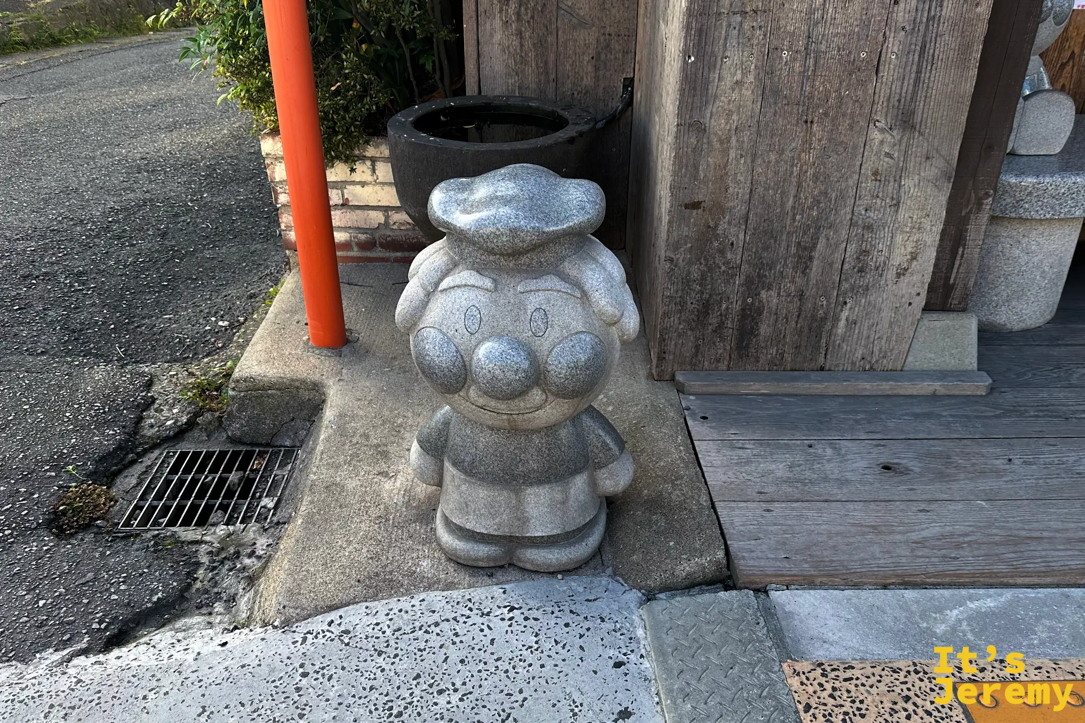 同一尊石雕麵包超人的正面特寫，圓圓的臉配上頭巾造型，站在石缸與木牆前