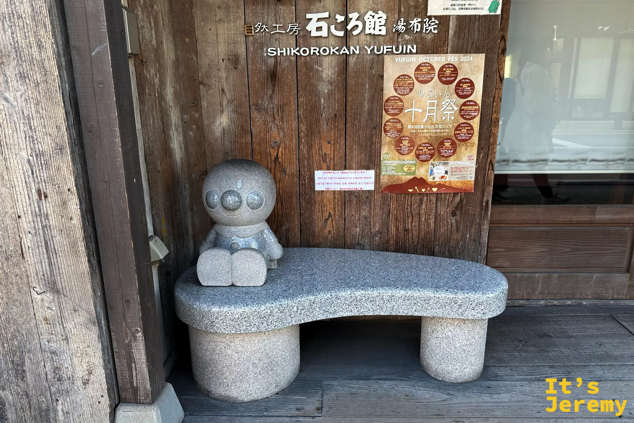 石ころ館 由布院門口的石凳上坐著一尊小小的石雕人物，背後是木造牆面與海報