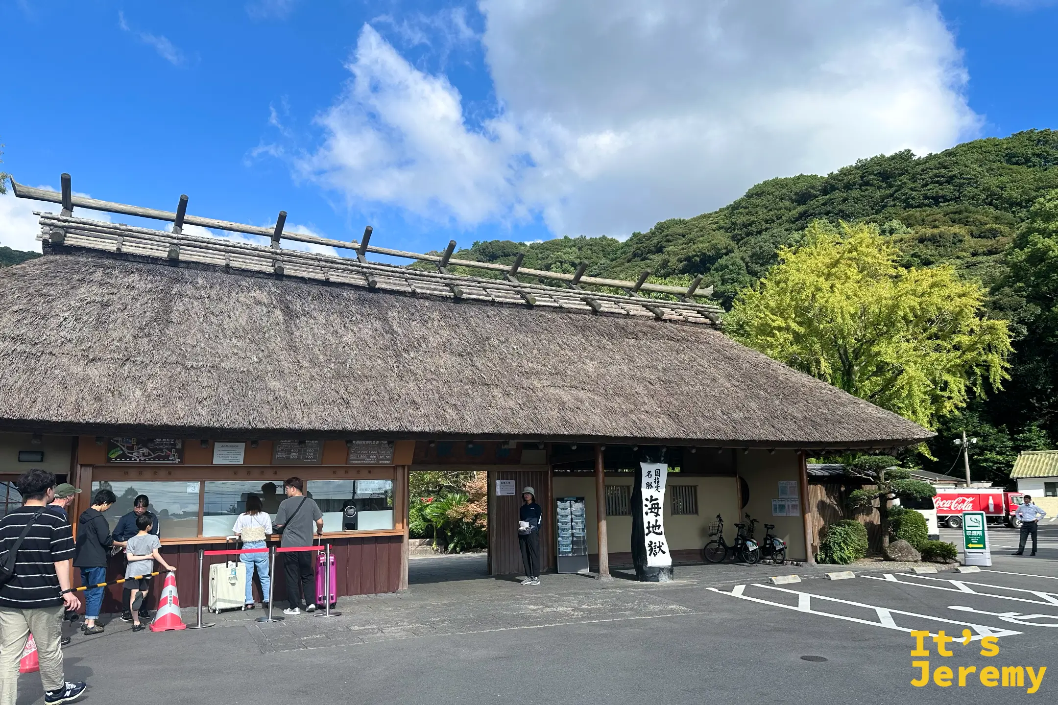 海地獄入口建築全景，遊客在售票窗口前排隊，後方是綠色山坡與藍天