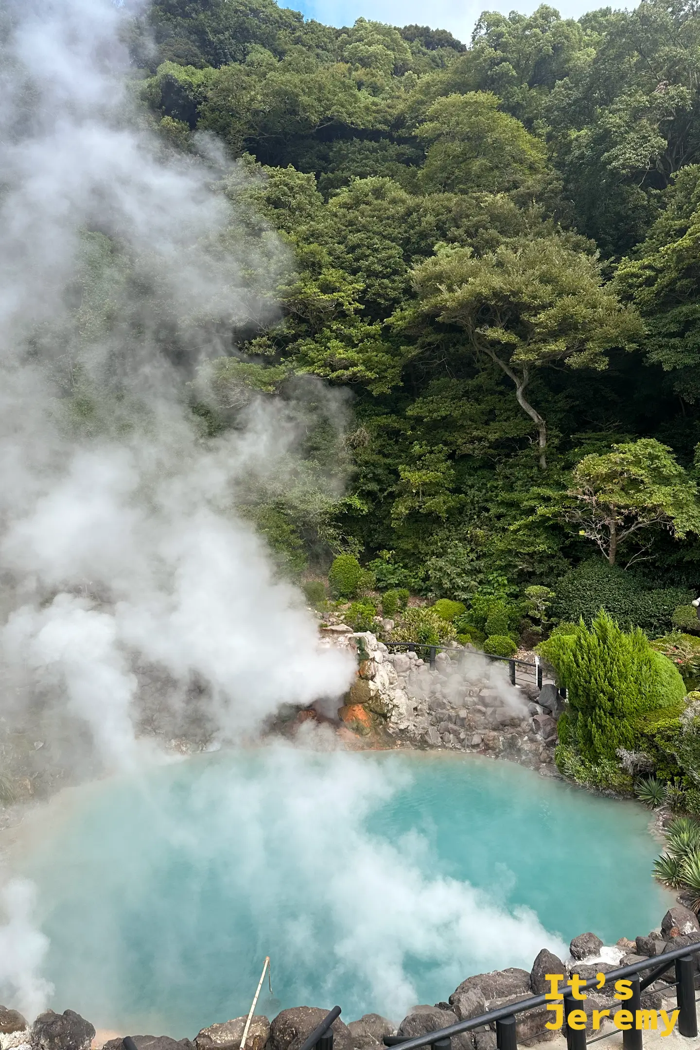 海地獄藍池與蒸氣的俯瞰角度，池畔以黑色欄杆與岩石圍起，四周是修剪整齊的庭園