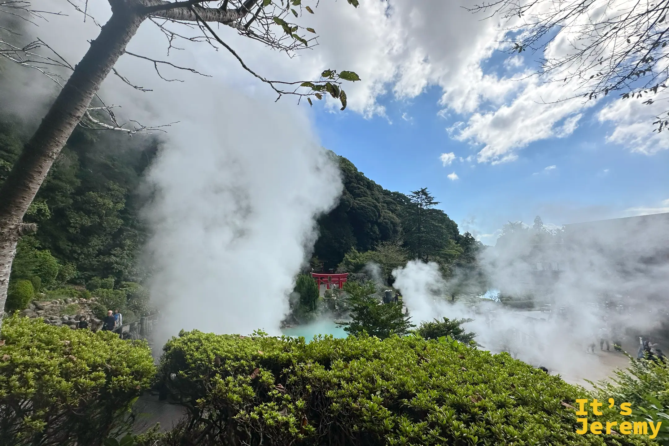 透過樹叢與灌木望向蒸氣中若隱若現的紅色鳥居，上方是藍天白雲