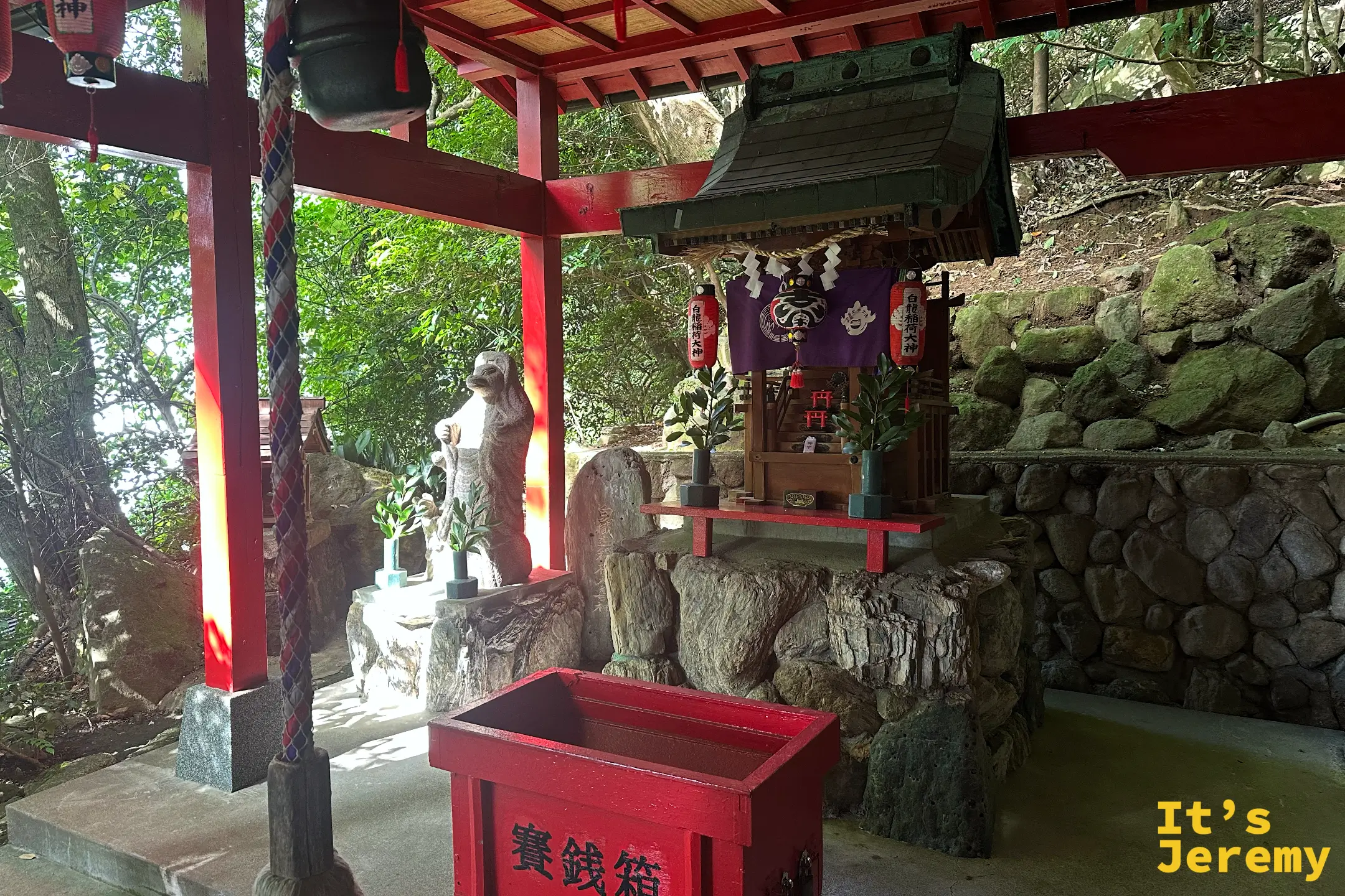 白龍稲荷大神的紅色小社，供著石狐、擺著紅色賽錢箱與紫色帳幔，背後是堆石護坡
