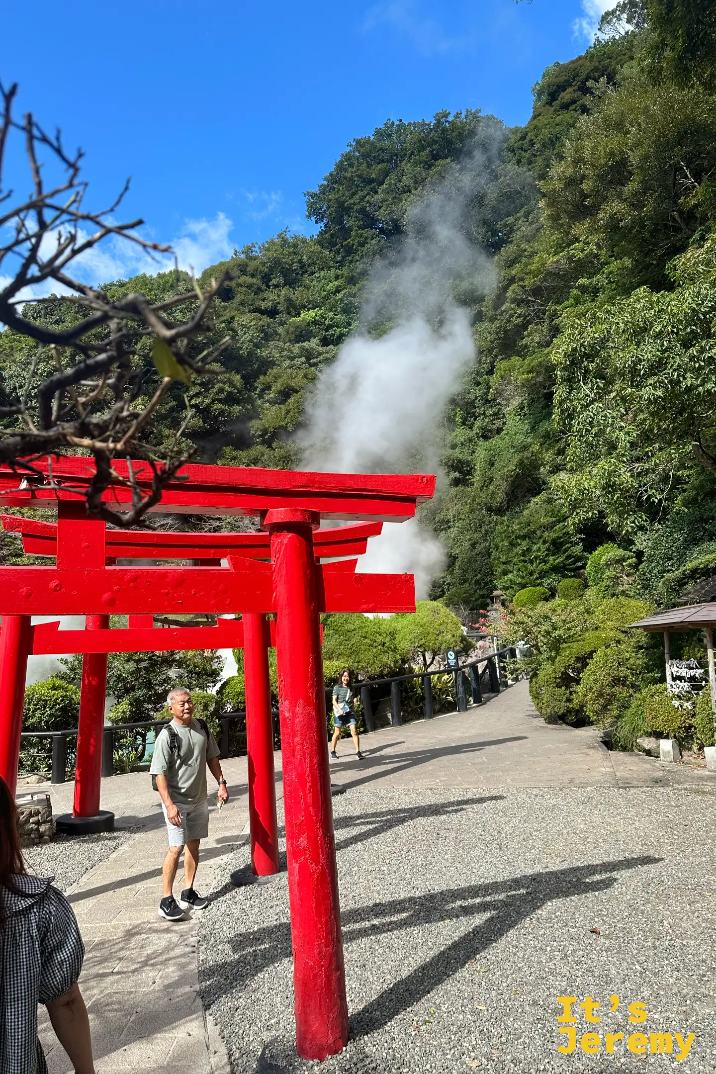 一座大型紅色鳥居橫跨在步道上，後方蒸氣柱不斷升起，遊客從鳥居下走過