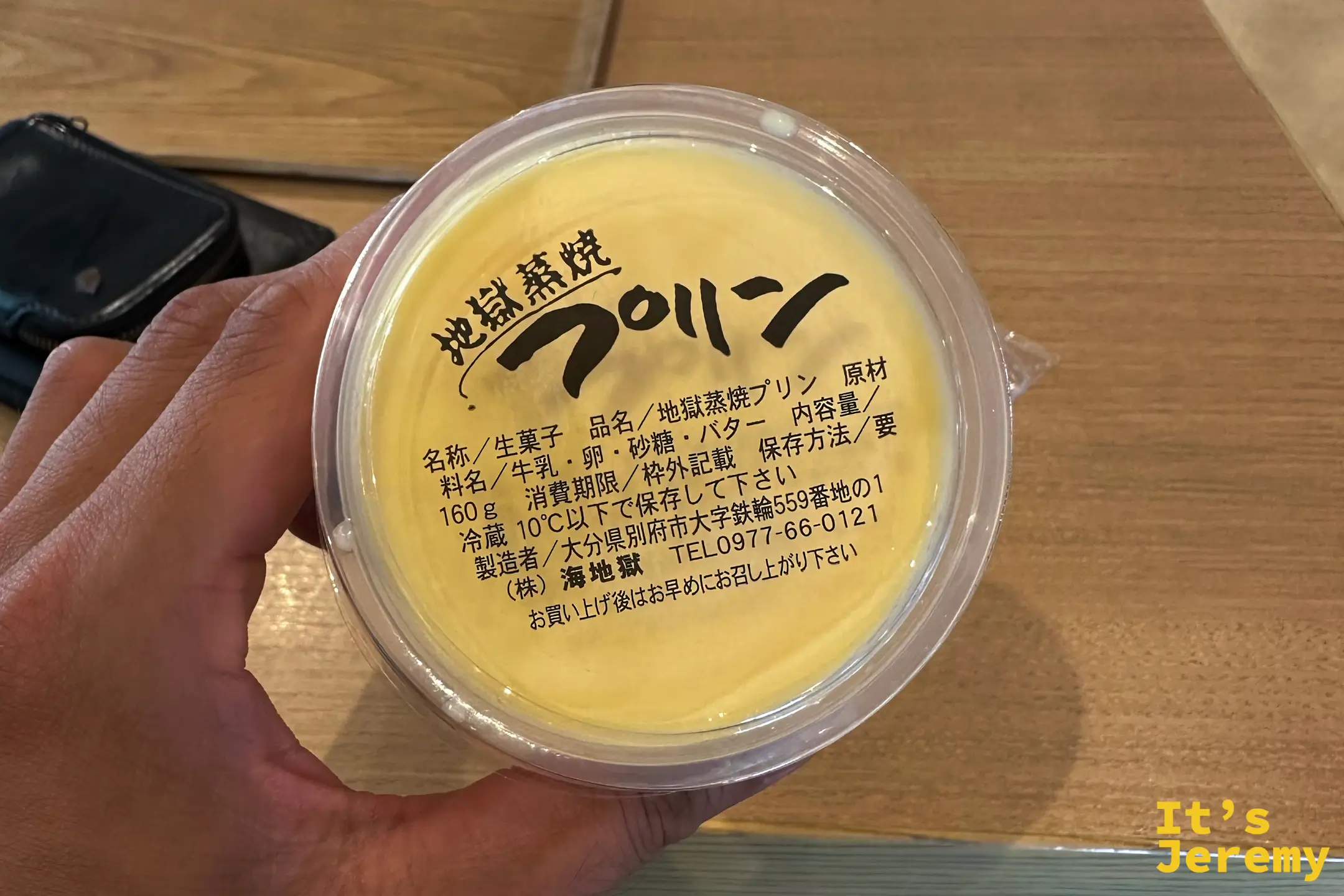手持一杯地獄蒸焼布丁的特寫，黃色布丁表面平滑，標籤標明產地為別府市鉄輪