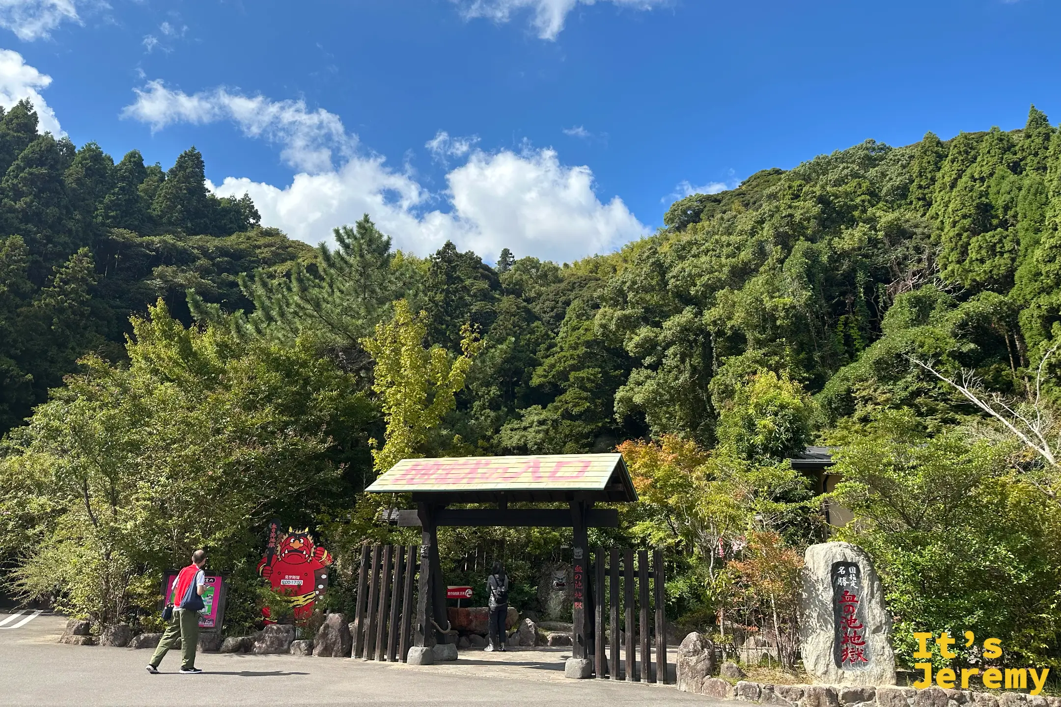血之池地獄的茅草小門入口，掛著黃底紅字「地獄の入口」，右側立著「国指定名勝 血の池地獄」石碑，旁邊有紅色鬼卡通立牌