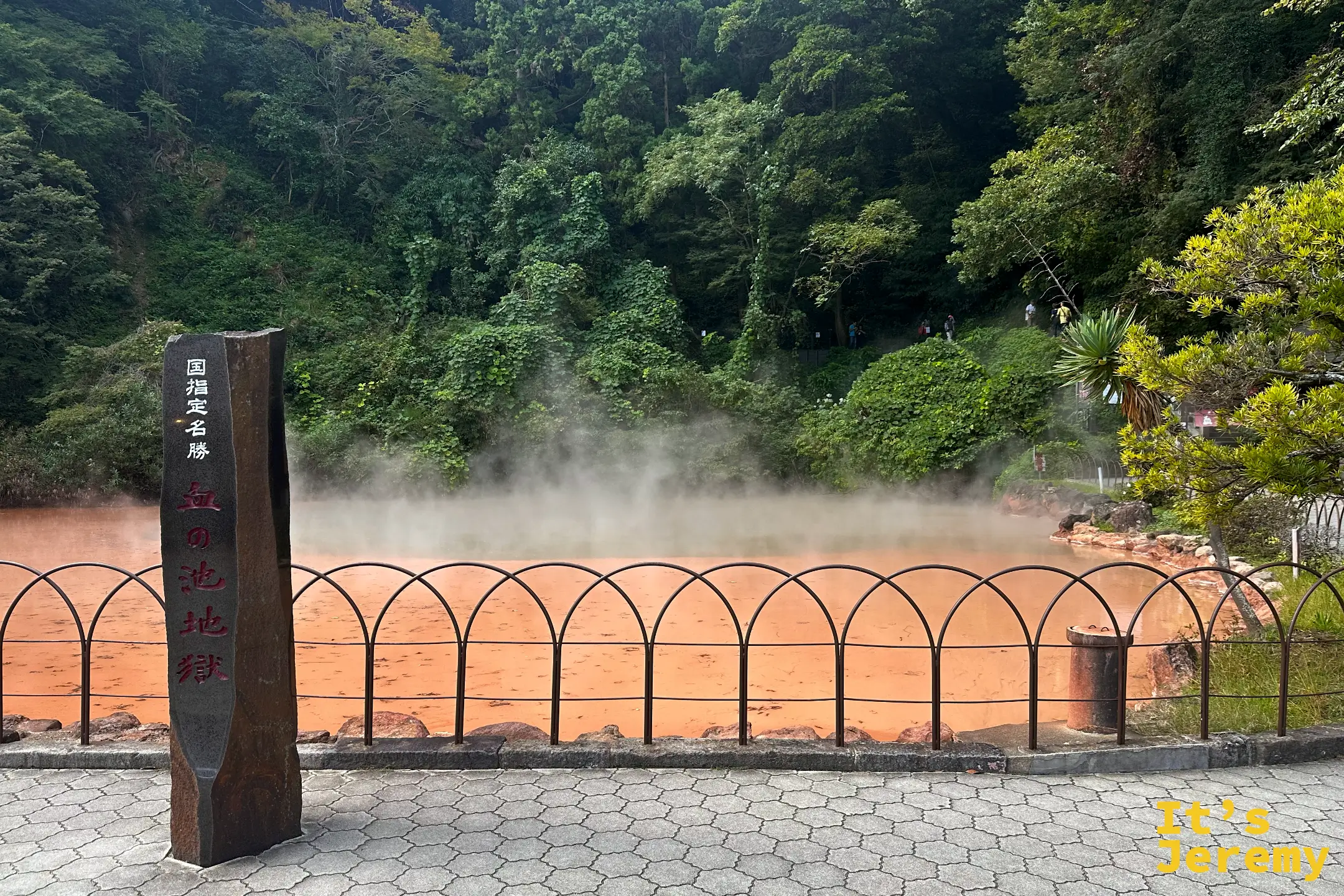 血之池地獄主池全景，橘紅色泥漿池被鐵欄杆圍起，立著「国指定名勝 血の池地獄」黑石碑，白煙升起