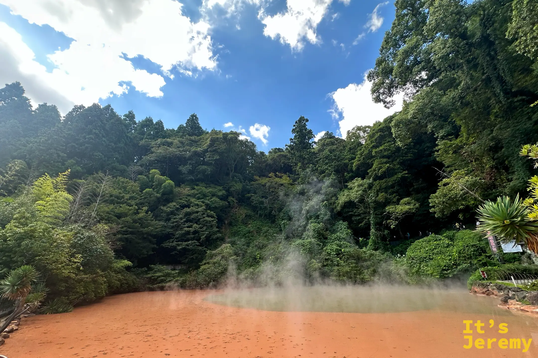 血之池地獄的橘紅色池水廣角，藍天白雲下四周被綠樹圍繞