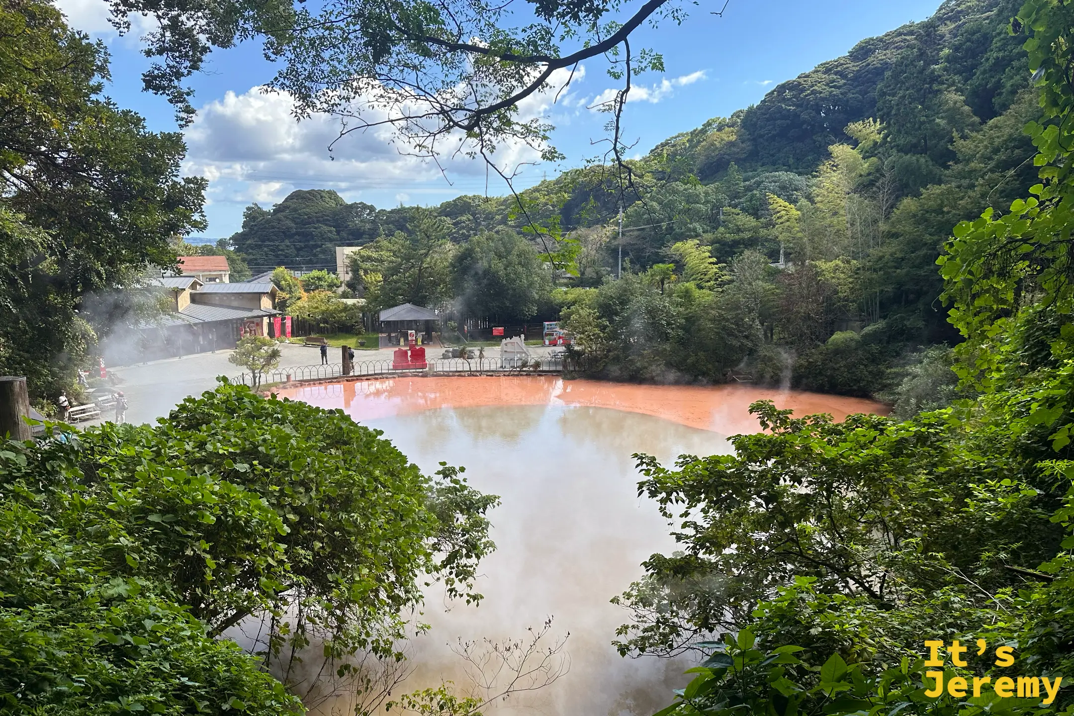 別府血之池地獄，一整池橘紅色的熱泥漿冒著白煙，池畔綠樹環繞