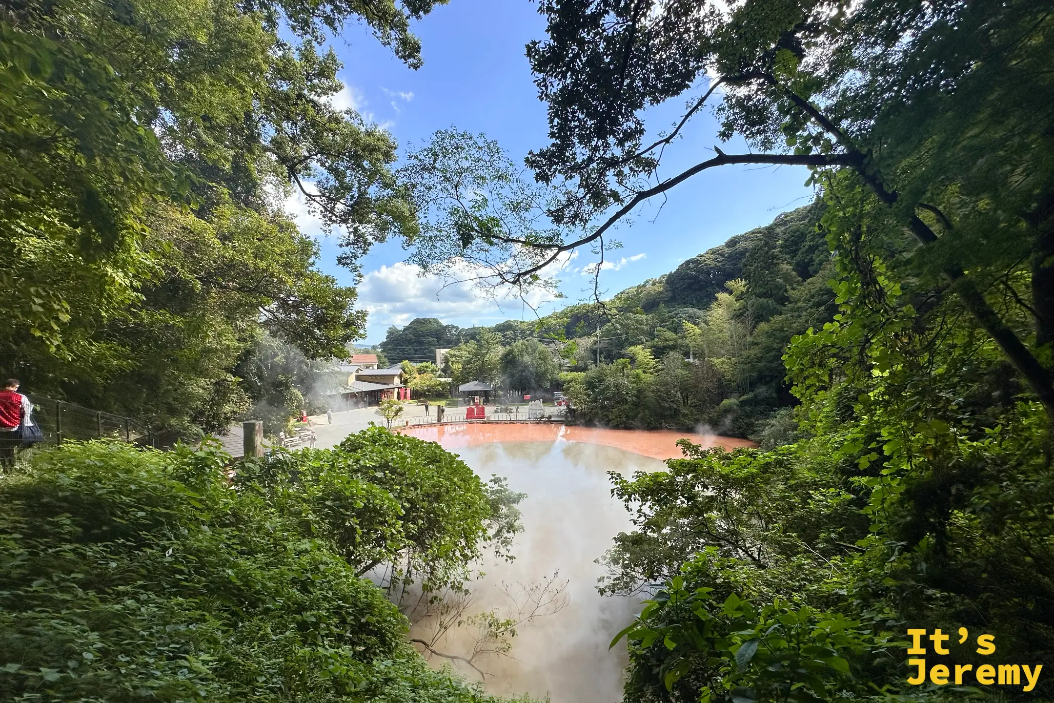 從樹叢間的高台俯瞰血之池地獄，橘紅池水與白煙，遠方可見賣店與紅色打卡看板