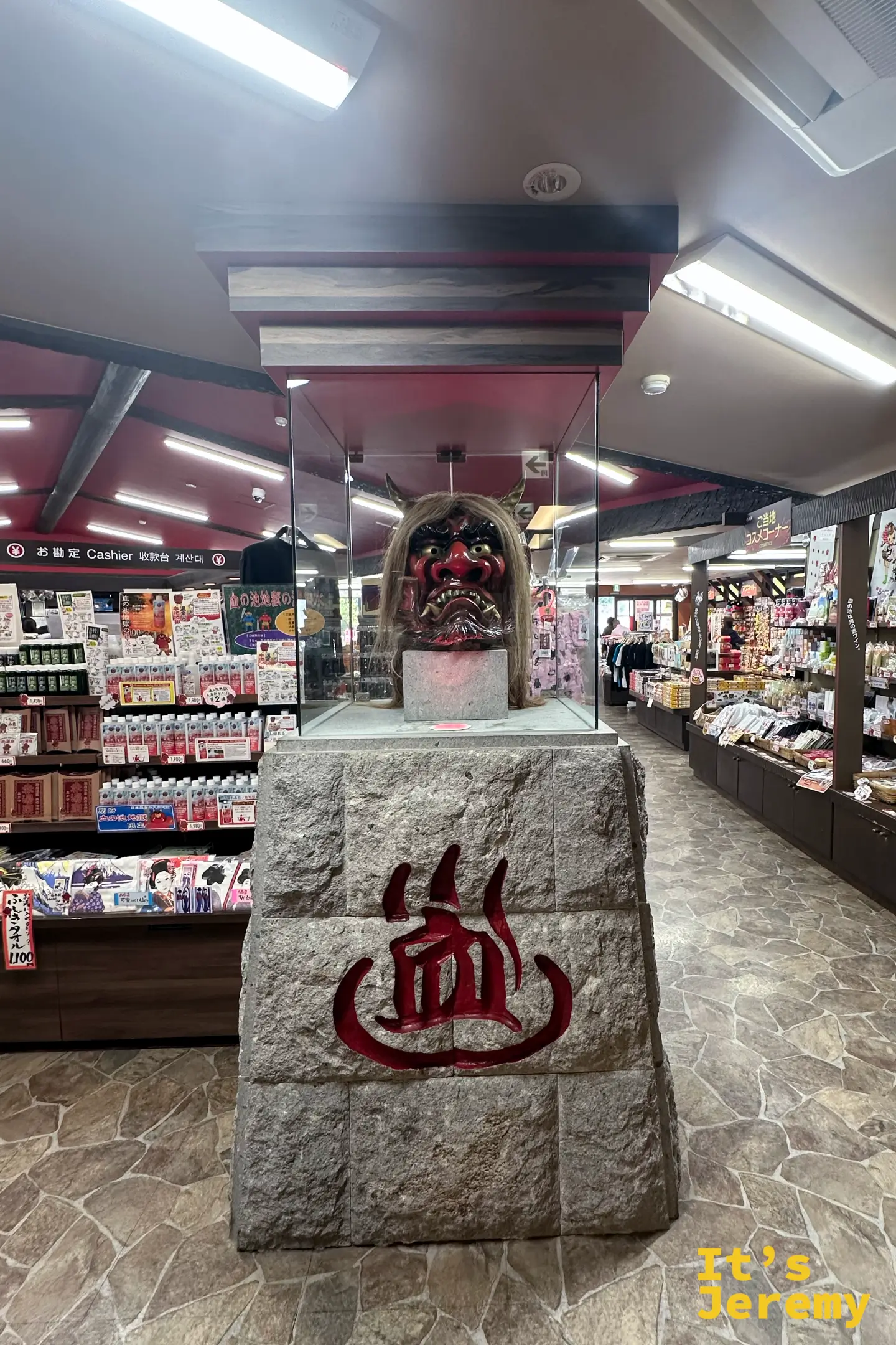 血之池地獄賣店內部，中央玻璃櫃裡展示著一張紅色鬼面具，石台上刻著紅色的「血」字，兩側是滿滿的伴手禮貨架