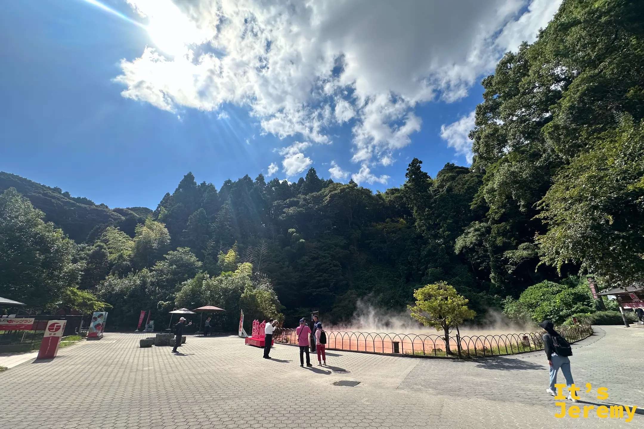 血之池地獄入口廣場全景，藍天下遊客在紅褐色池邊拍照，鐵欄杆沿著池畔延伸