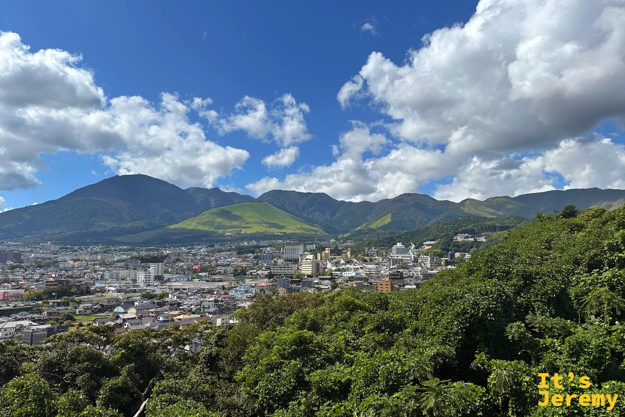從湯煙展望台望向別府市街與鶴見岳連峰，藍天白雲下綠色草山格外醒目