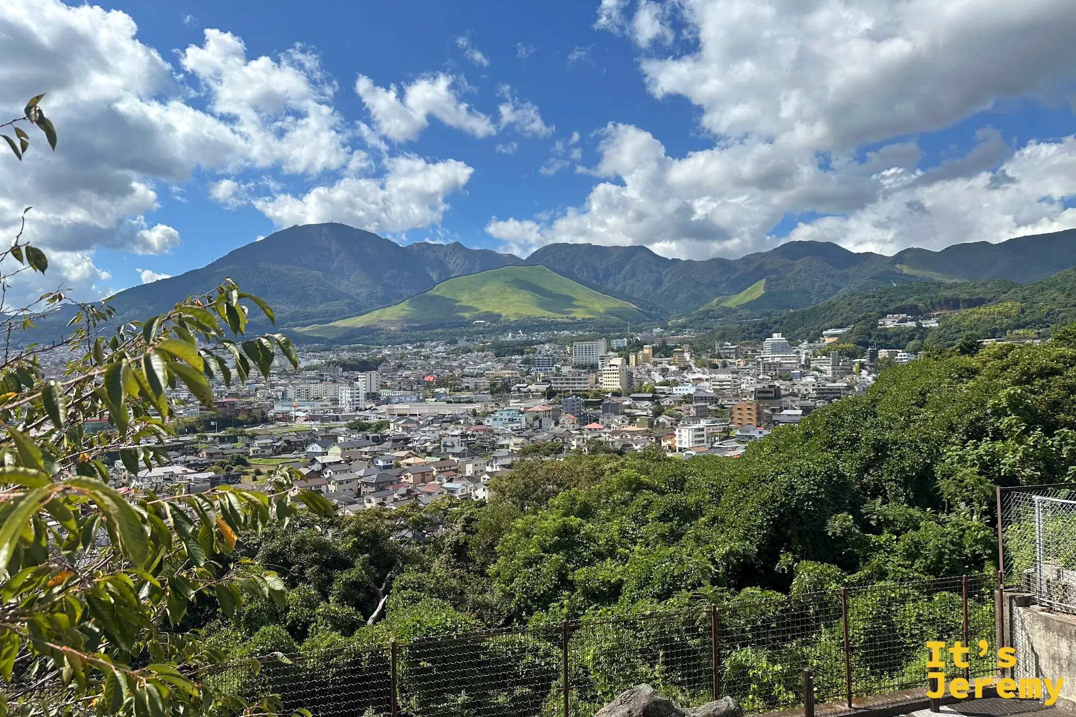 從樹葉框景望出去的別府市街與鶴見岳連峰，前景有觀景台的鐵絲網欄杆