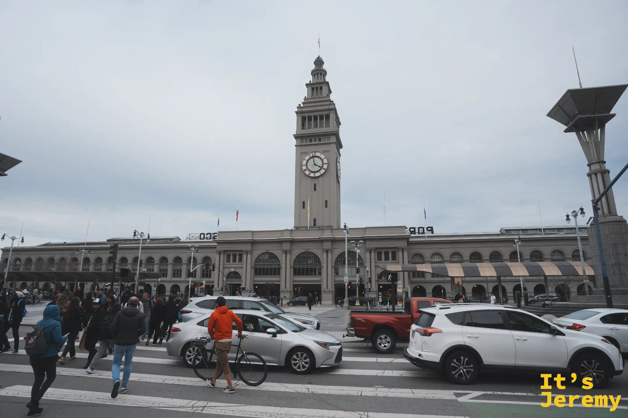 從濱海大道對街望向渡輪大廈，馬路上車來車往，行人正穿越斑馬線，鐘塔立在建築中央