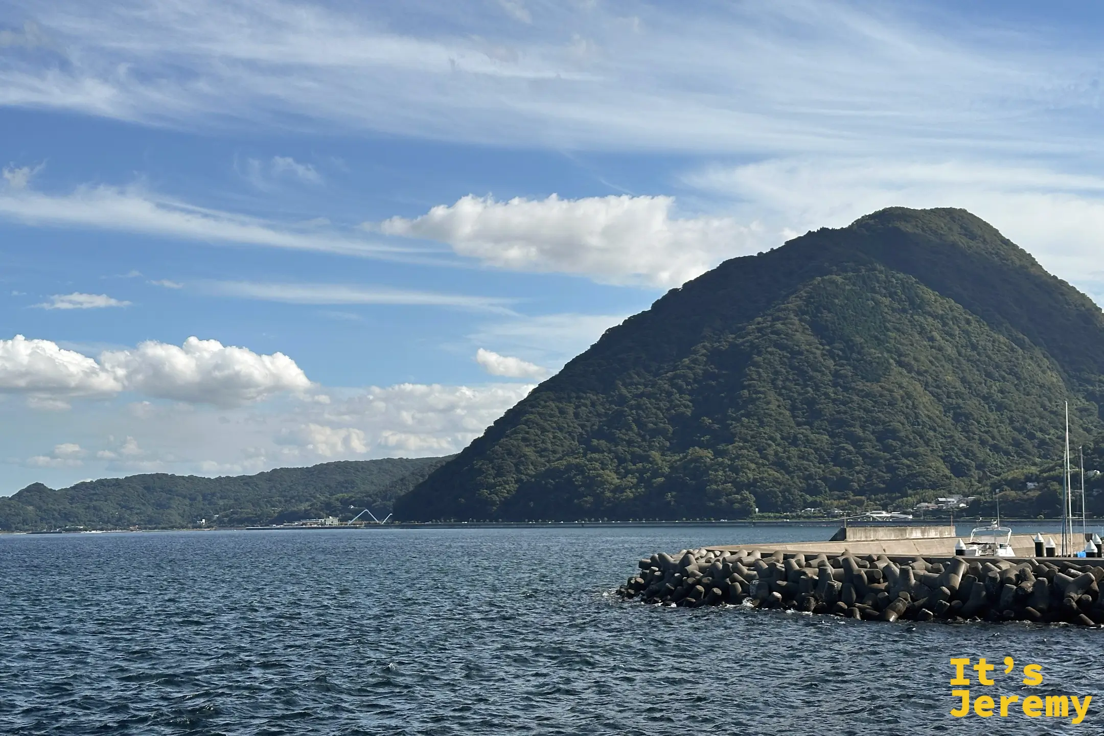 別府灣海面上，一座圓錐狀的綠色小山立在海的另一頭，前方有防波堤