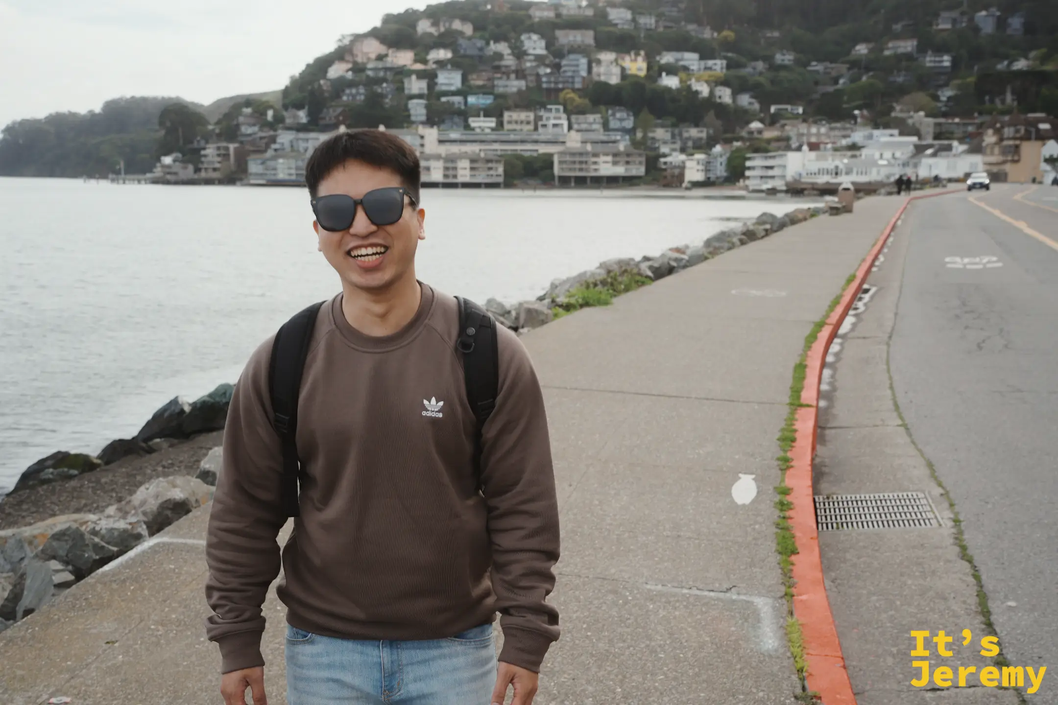 Jeremy 戴著墨鏡在海濱步道上笑著，背後是索薩利托山坡上層層疊疊的房子