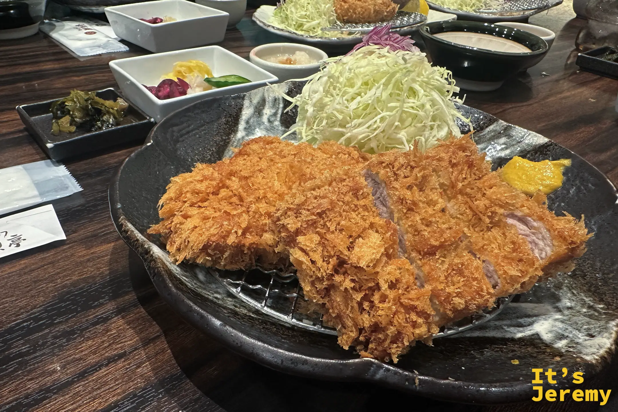 厚切炸豬排的側面特寫，酥脆炸衣包著厚實的豬肉，後方是高麗菜絲與漬物