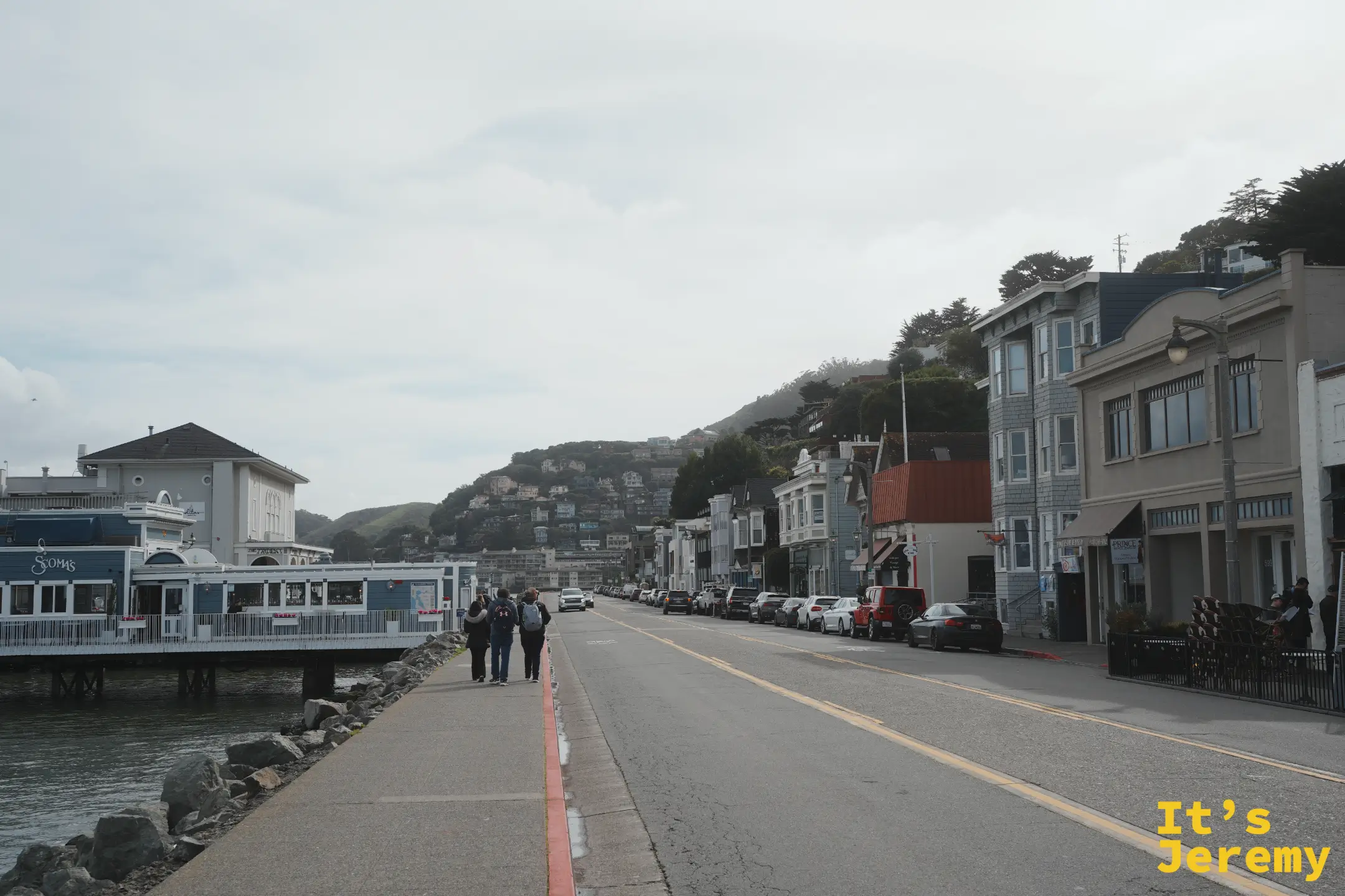 從海濱步道望向小鎮街道，左側是水上的餐廳建築，右側是一整排臨街店家與背後的山丘