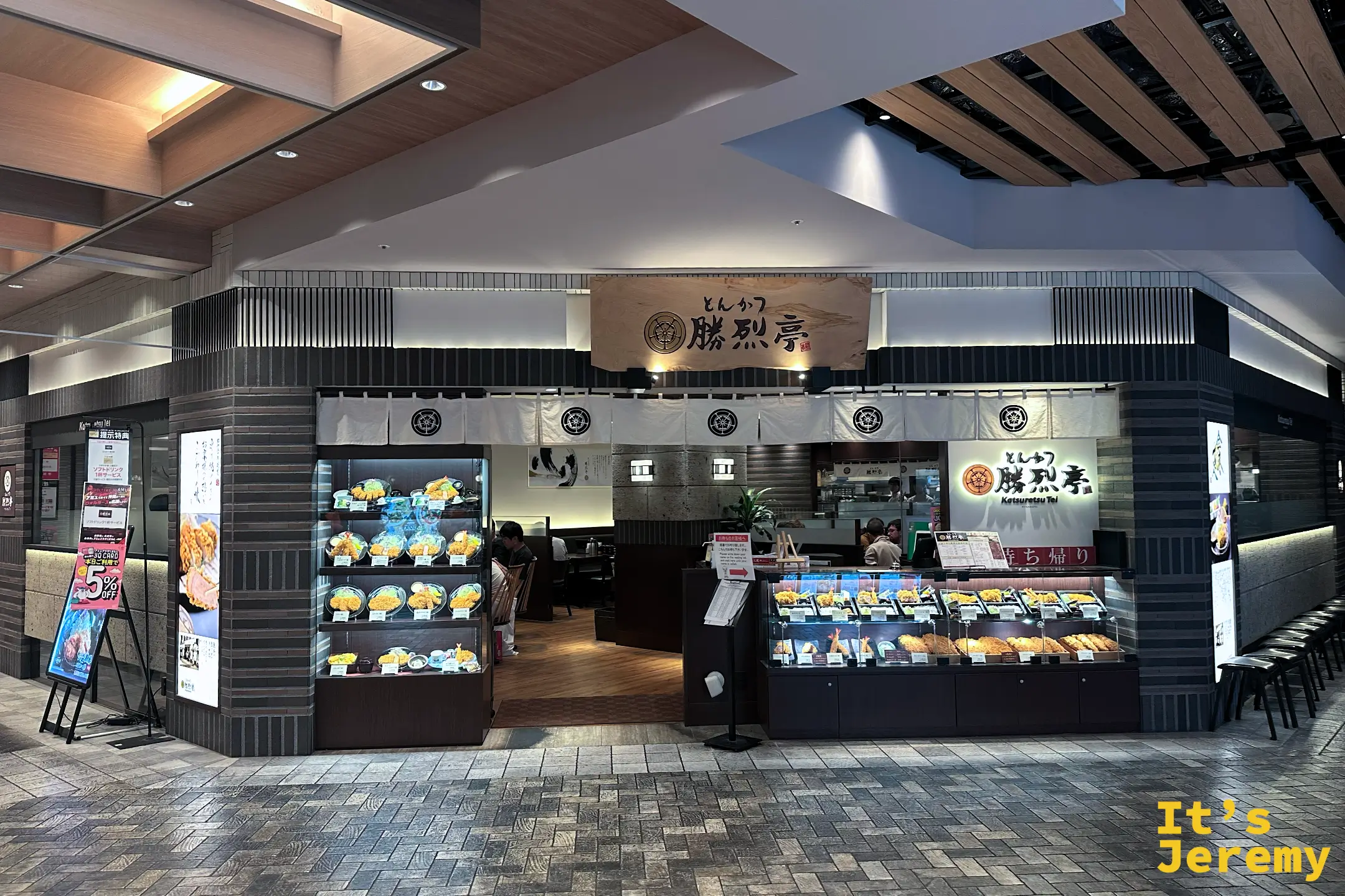 勝烈亭在 AMU PLAZA 熊本的店面，木質招牌寫著「とんかつ 勝烈亭 Katsuretsu Tei」，門口有食物模型展示櫃與外帶櫃台