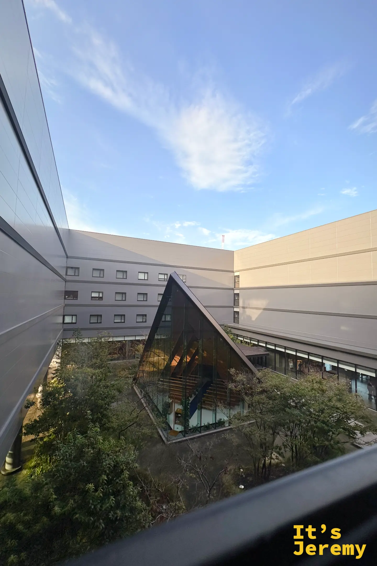 清晨從熊本旅館高處往中庭望，一座三角形玻璃屋建築藏在綠樹之間，藍天無雲