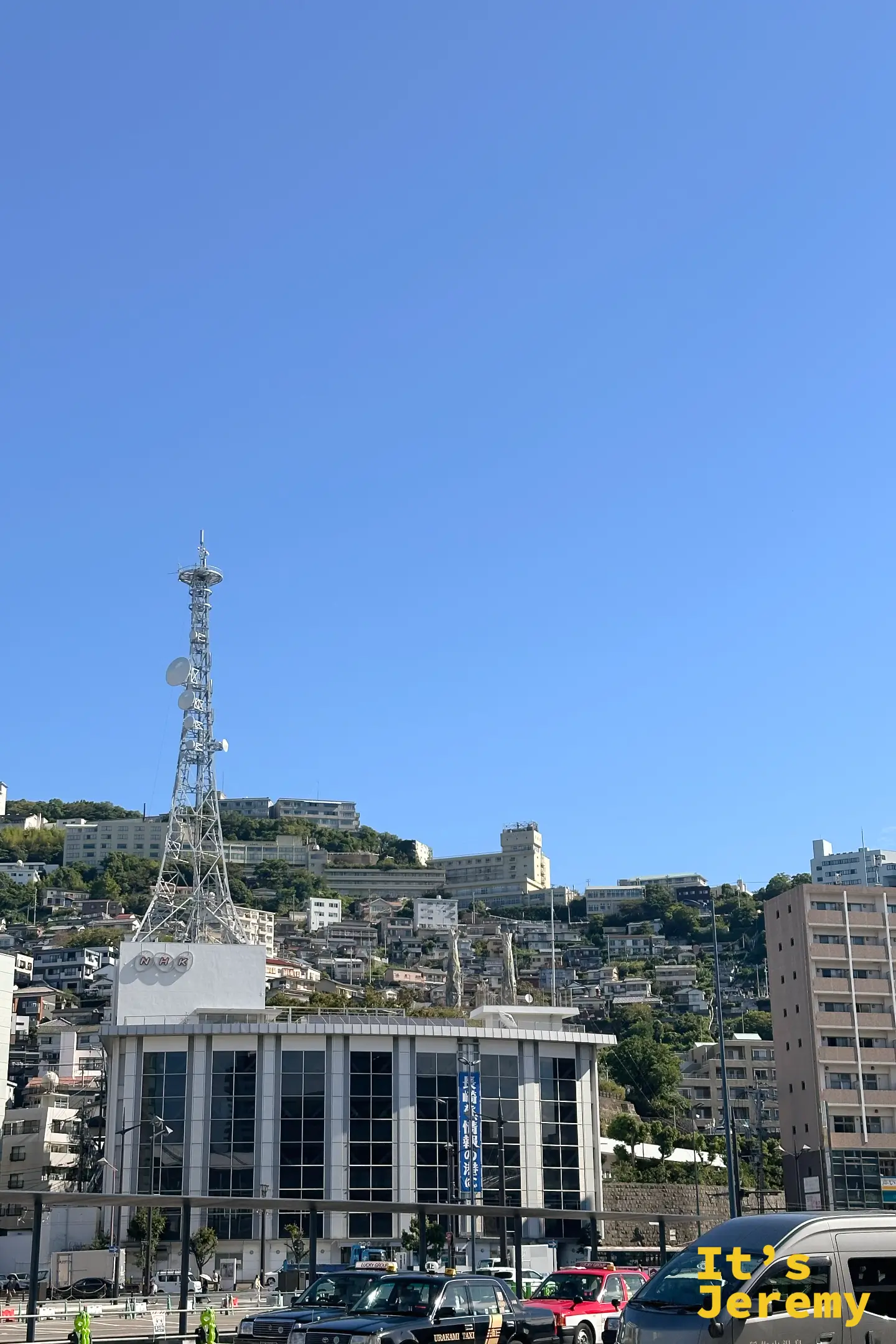 長崎車站前望向山坡，密集的住宅沿山而建，山頭立著高聳的電波塔，前方有計程車
