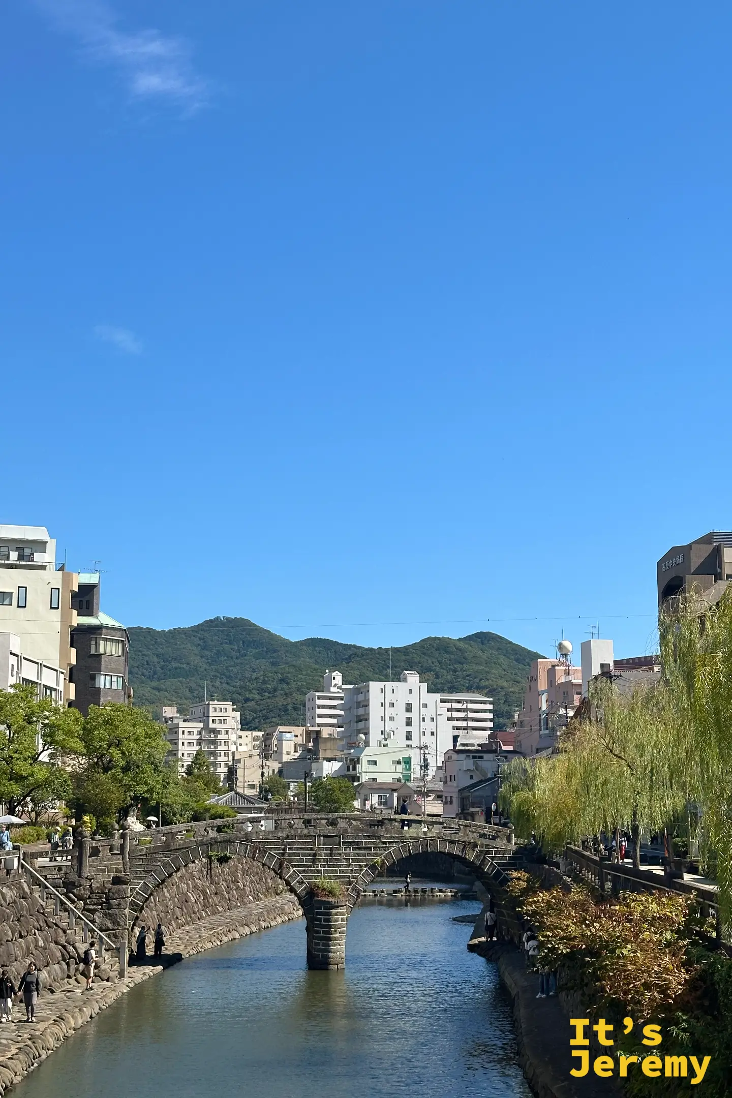 從遠處望向眼鏡橋，石橋低低地橫在中島川上，後方是長崎的山城與藍天