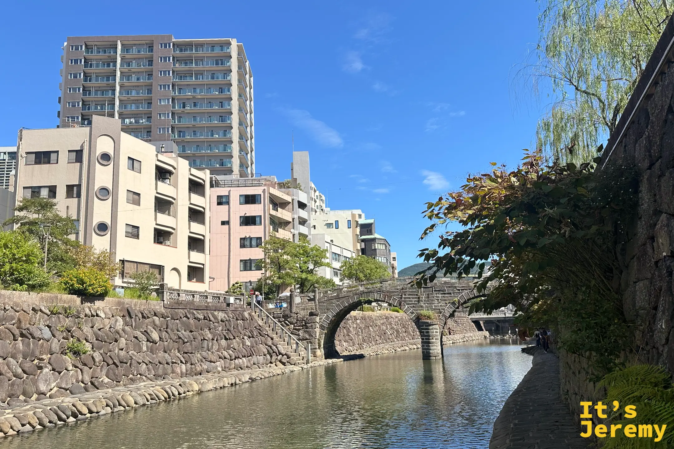 中島川沿岸的石砌護岸與柳樹，左岸有一棟圓窗老建築，河道兩旁是長崎市街