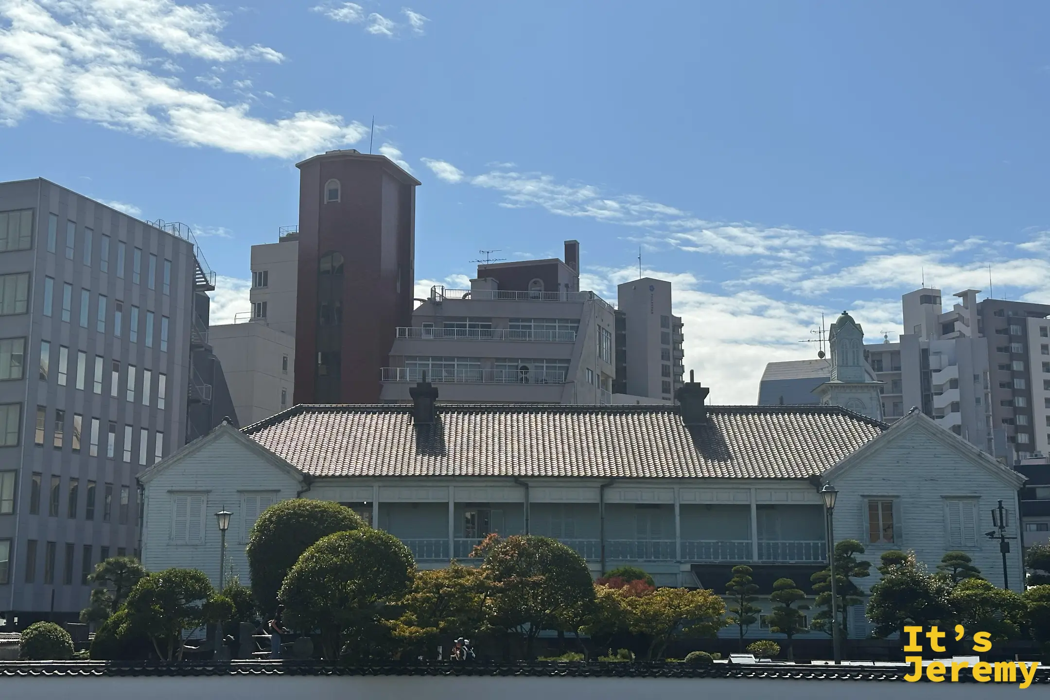 隔著街道看出島，成排的淺綠色西洋商館建築與修剪整齊的庭園樹，後方是現代長崎的高樓