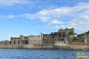 海上浮現的軍艦島側影，高聳護岸後方是層層疊起的水泥廢墟