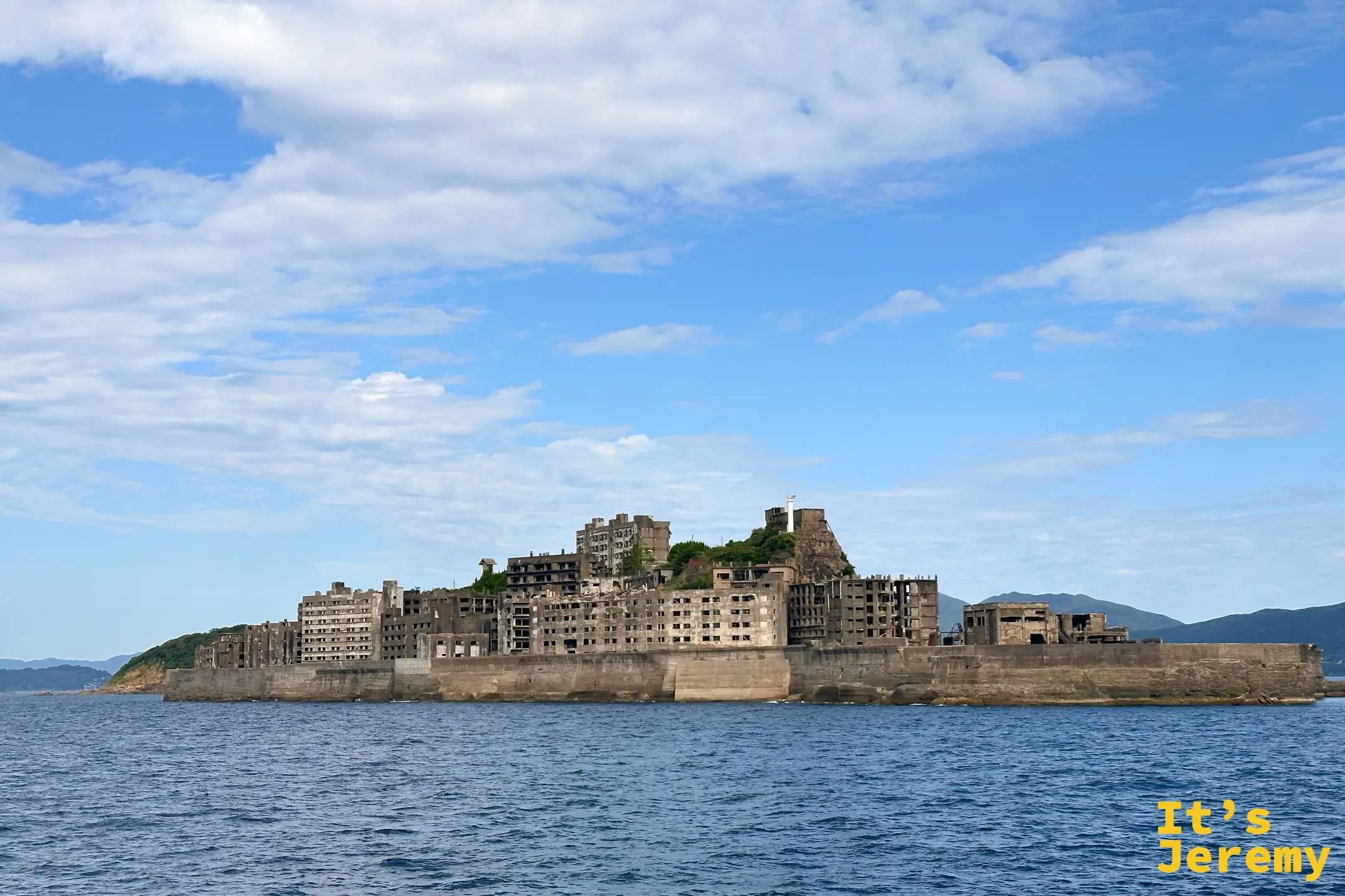 海上遠望軍艦島全貌，長長的護岸包住整座島，山頂有燈塔