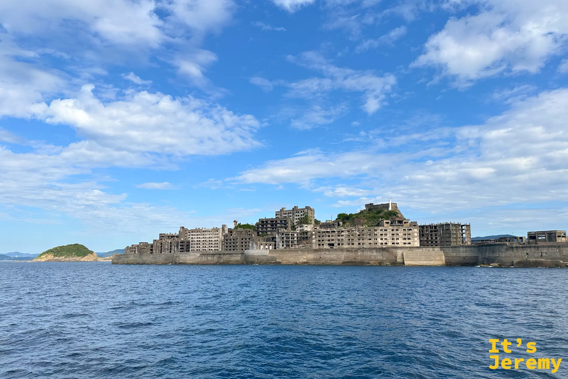海上望向端島，長長的護岸包住整座島，山頂有燈塔，左側是一座綠色小島