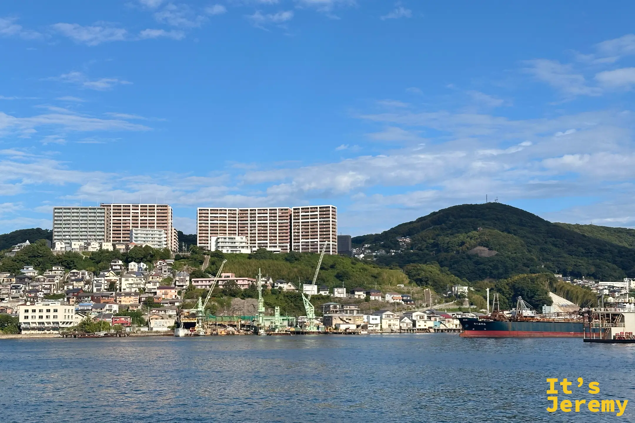 長崎港岸邊沿山而建的住宅，山頂矗立著大型集合住宅，岸邊有造船廠起重機與一艘紅色船身的貨船