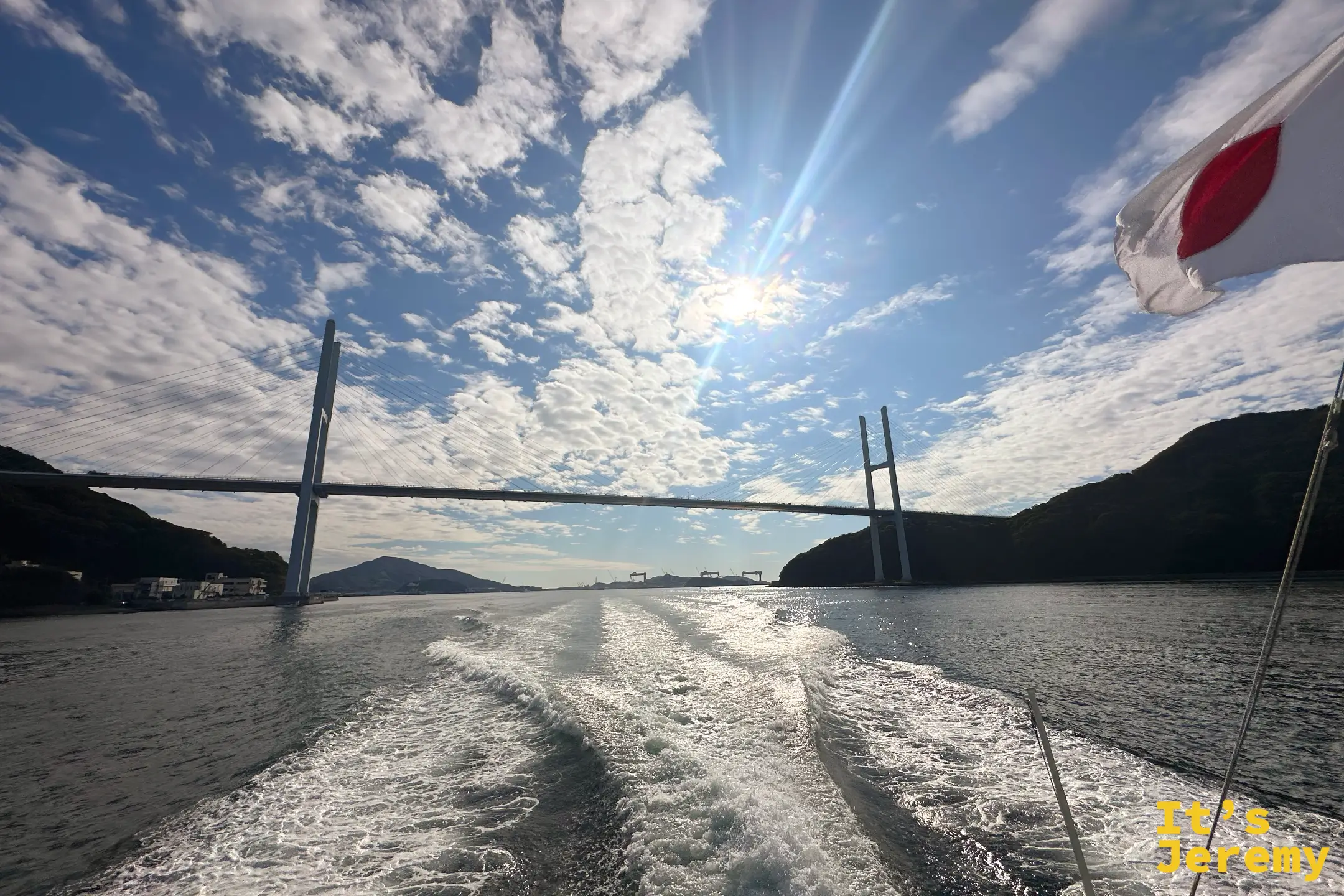 回程逆光的女神大橋橫跨海面，陽光在雲間灑成放射狀，右上角是船尾的日本國旗
