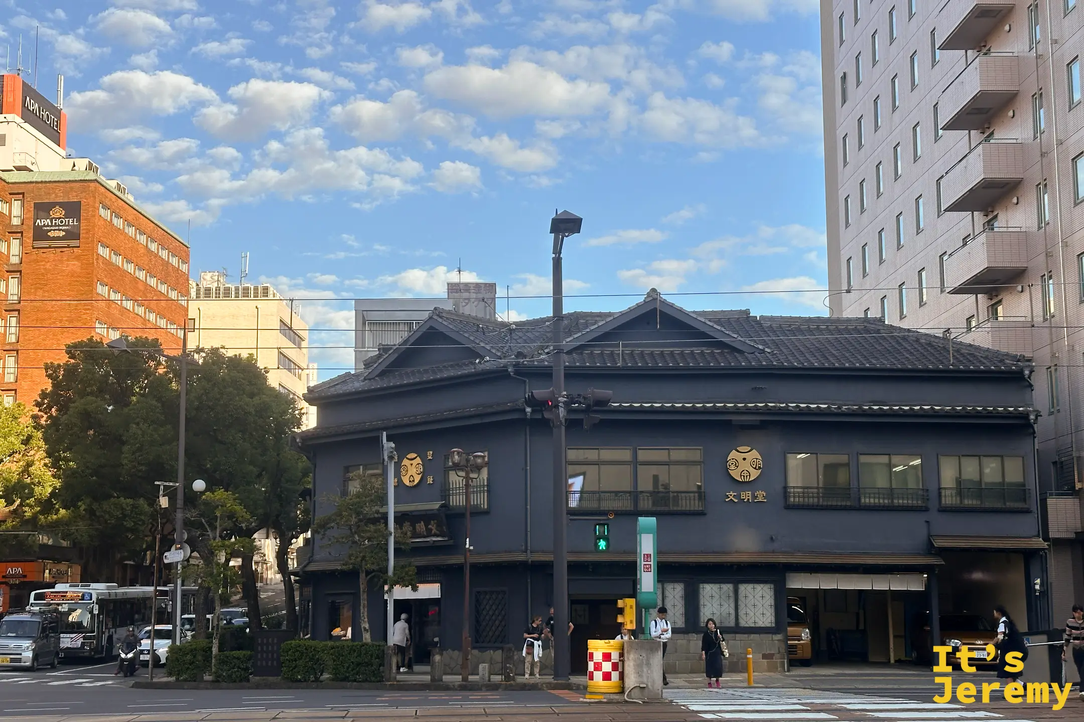 文明堂総本店外觀，深色木造老屋配上圓形金色堂號，立在長崎市區街角，傍晚天色