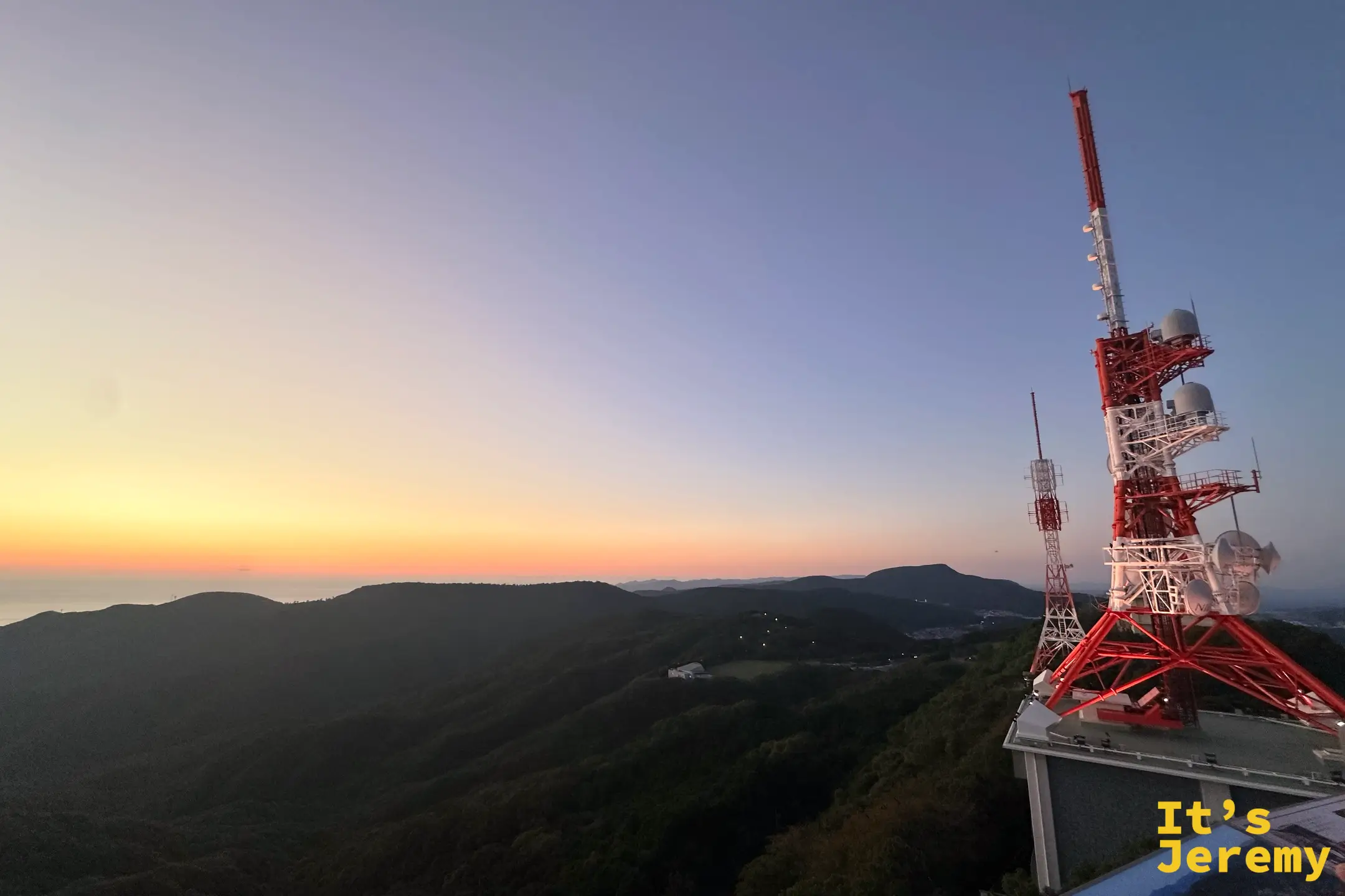 夕陽餘暉中的稲佐山頂紅白轉播鐵塔，遠處是漸暗的山景