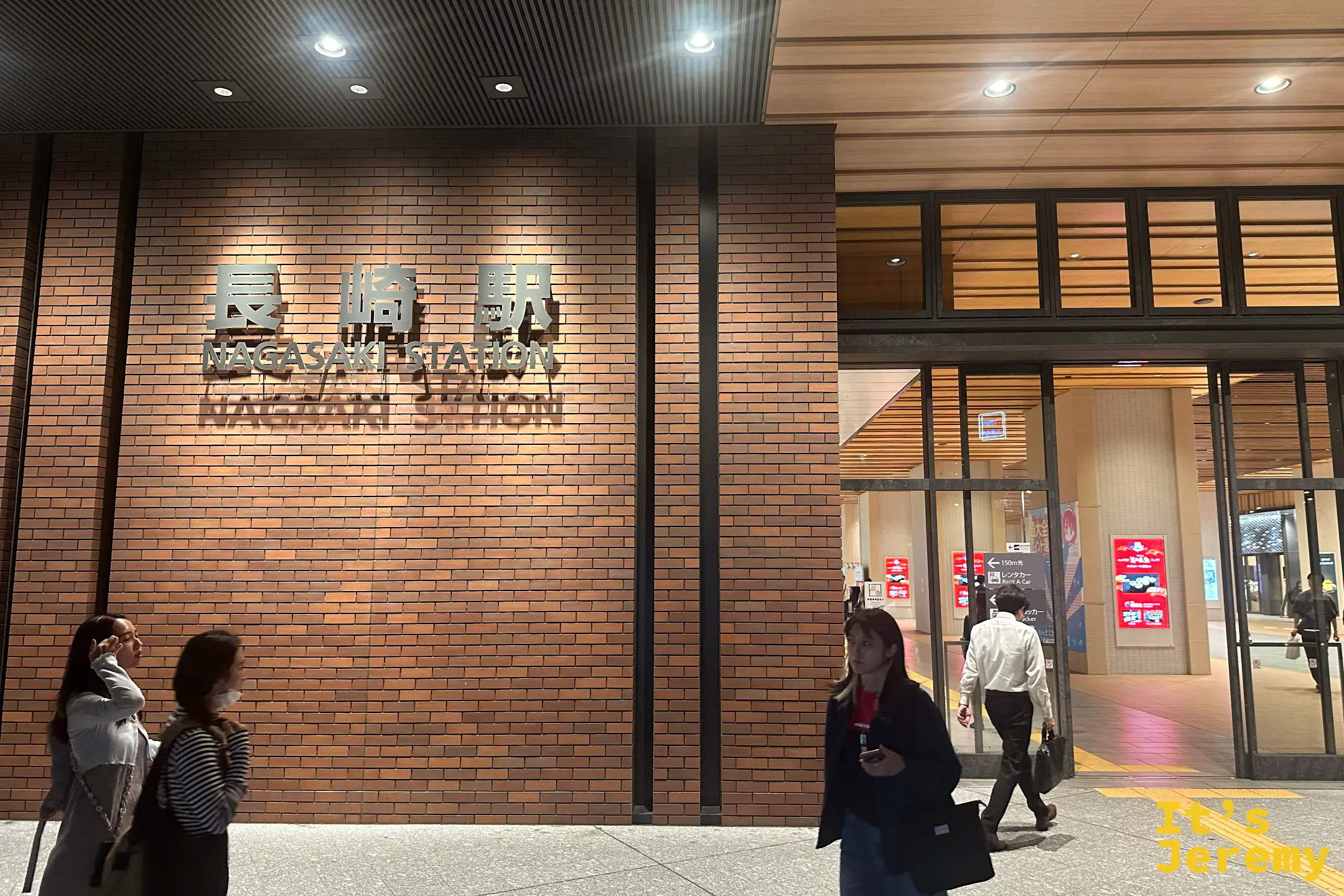 紅磚牆上「長崎駅 NAGASAKI STATION」的金屬字樣特寫，夜裡有旅客經過