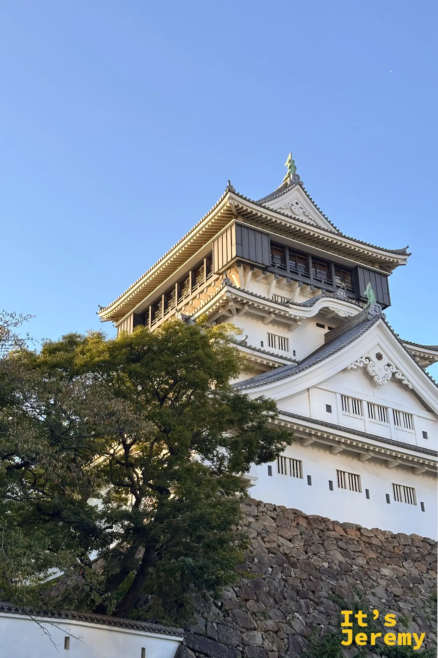 福岡北九州小倉城天守閣直式近拍，透過翠綠樹木作為前景，展現日本名城與自然交織的四季美感