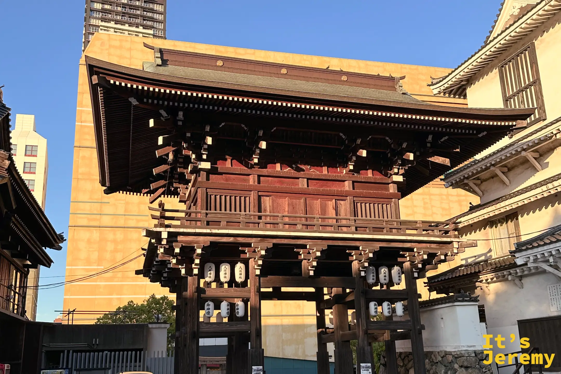從左側斜向視角拍攝北九州小倉八阪神社的東楼門，展現朱紅樓門與周邊林木交織的立體建築美感