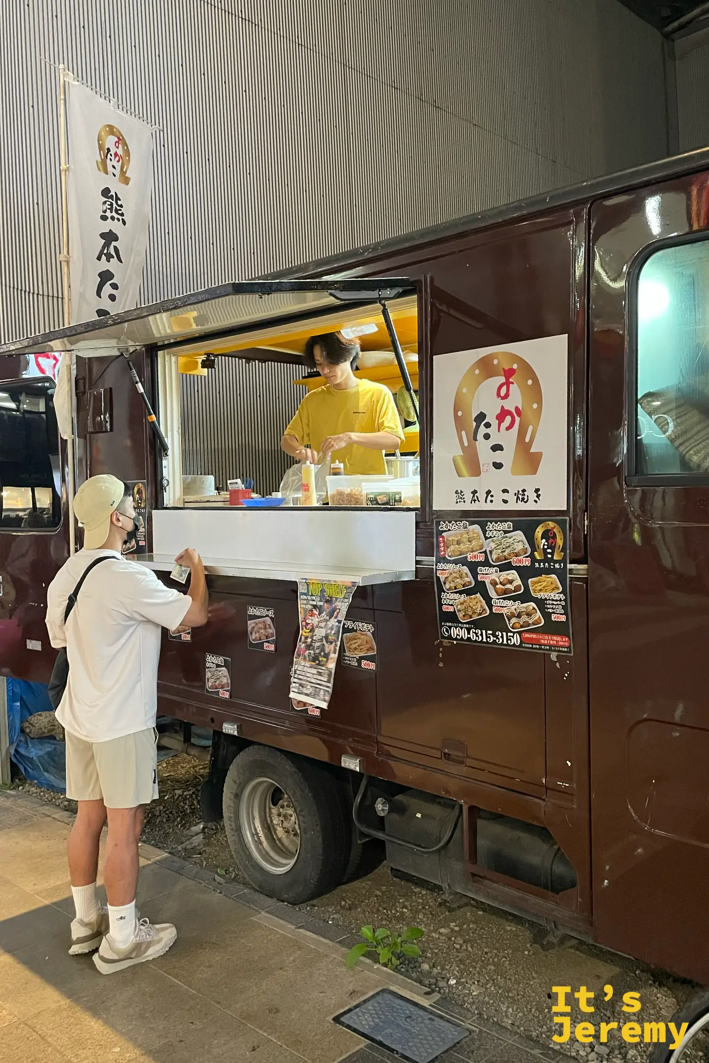 深棕色的「よかたこ」章魚燒餐車停在騎樓下，穿黃衣的老闆在窗口現做，車身貼著菜單，一名戴帽子的客人正遞錢點餐