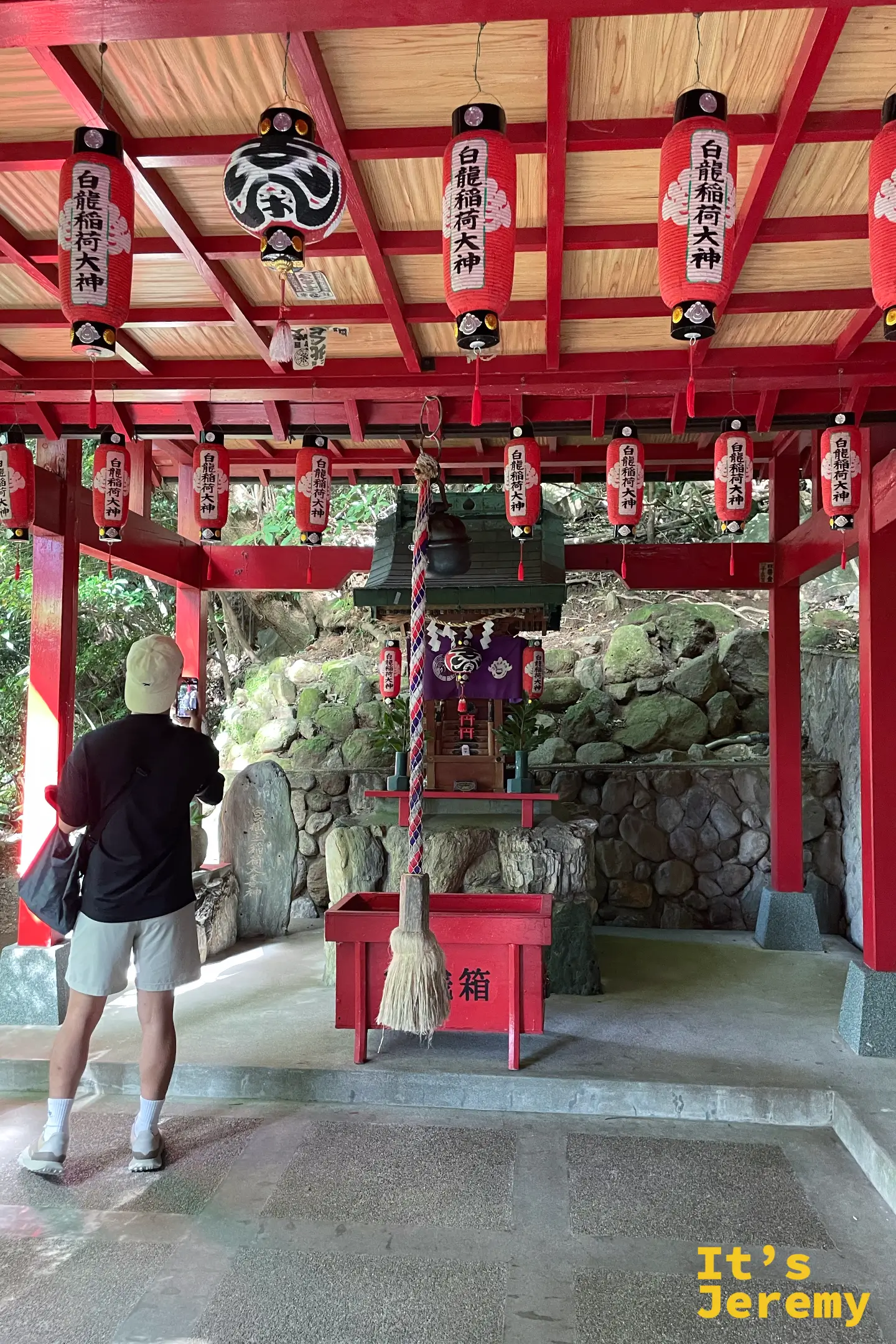 一名遊客舉起手機拍攝白龍稲荷大神，成排紅燈籠寫著「白龍稲荷大神」，中央垂著鈴繩與賽錢箱