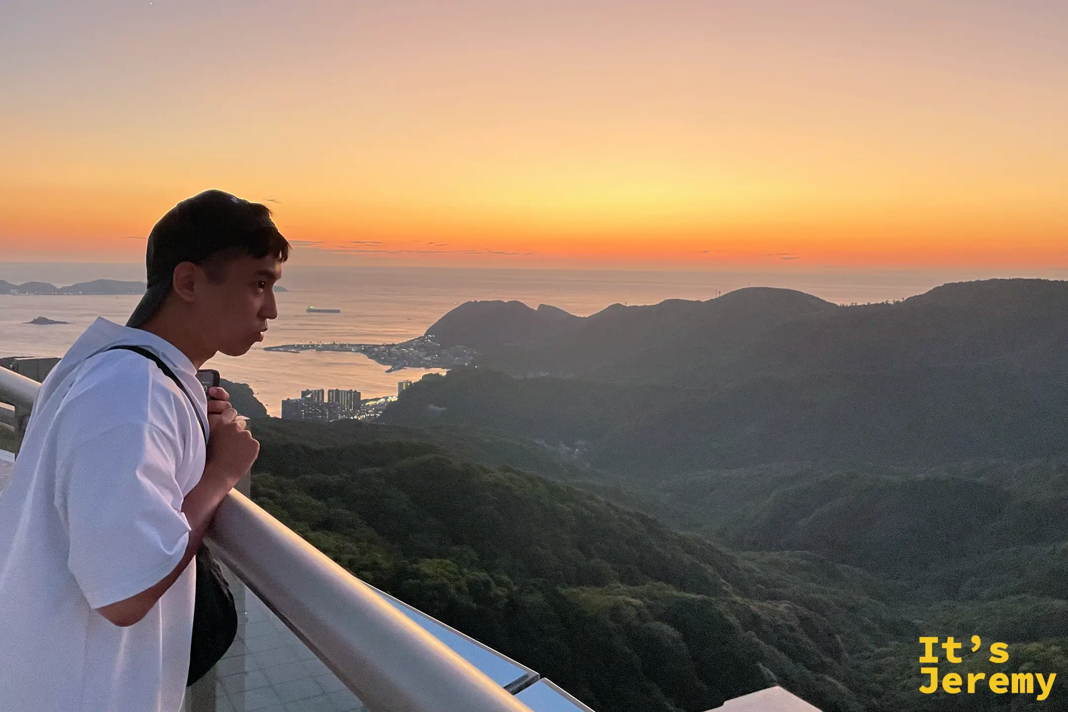 夕陽下的稲佐山山頂，一名遊客倚著欄杆望向橘紅色的海與山