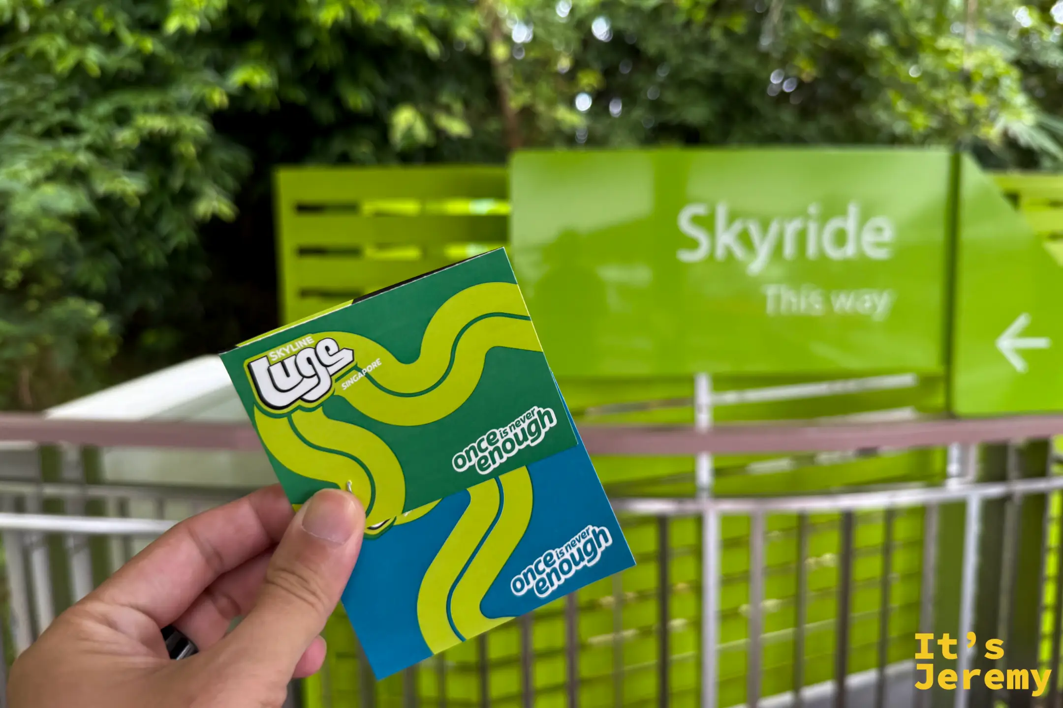 遊客手拿兩張黃綠條紋的 Skyline Luge 實體門票，背景為 Skyride 空中吊椅的綠色指標路牌