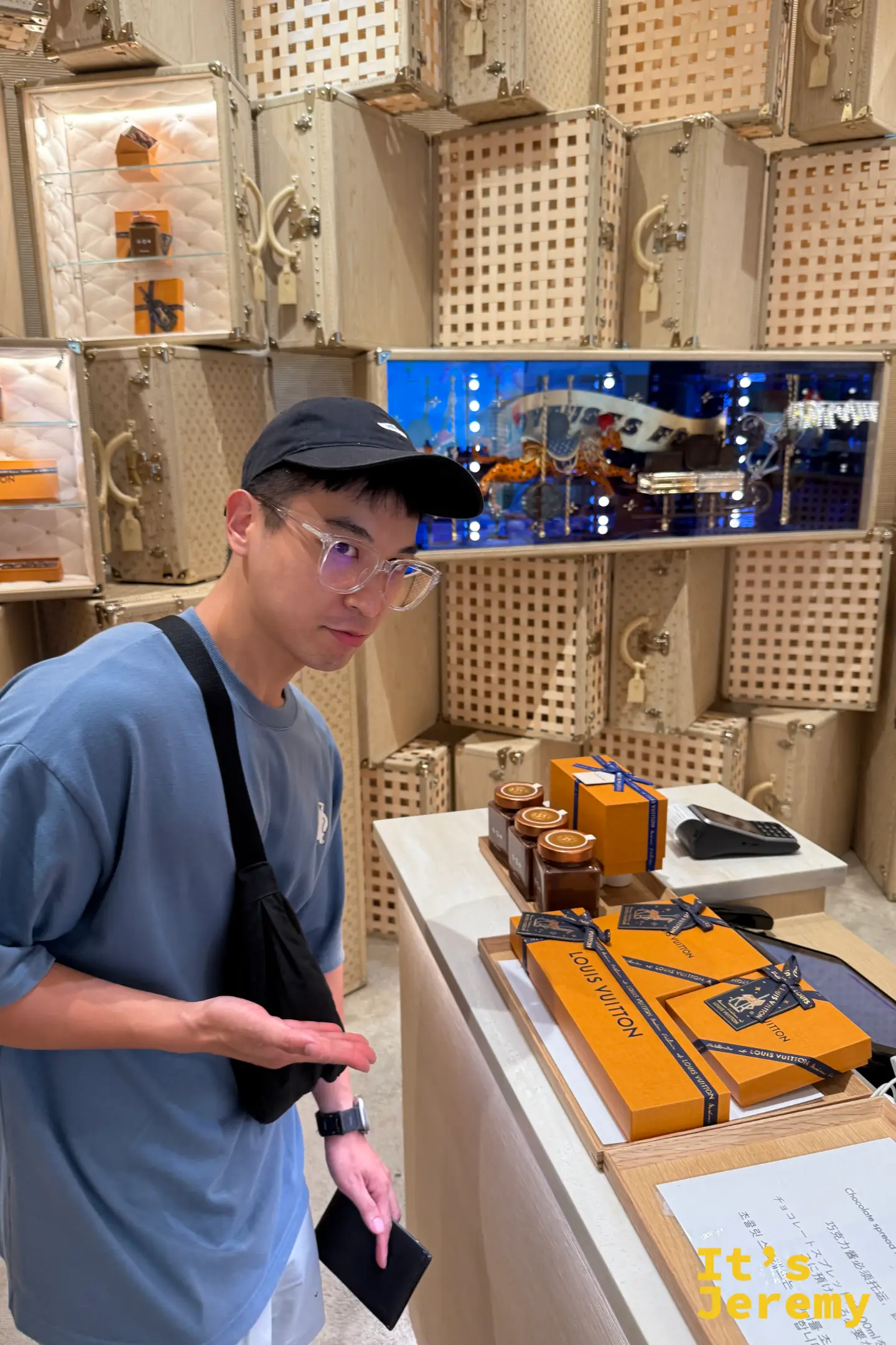 遊客近距離端詳 Louis Vuitton 與 Maxime Frédéric 聯名的限定巧克力系列,背景是店內堆疊如藝術裝置般的路易威登經典皮箱與木質方格壁面