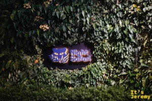 夜間野生動物園入口的紫色貓頭鷹霓虹招牌，融入綠意盎然的植物牆