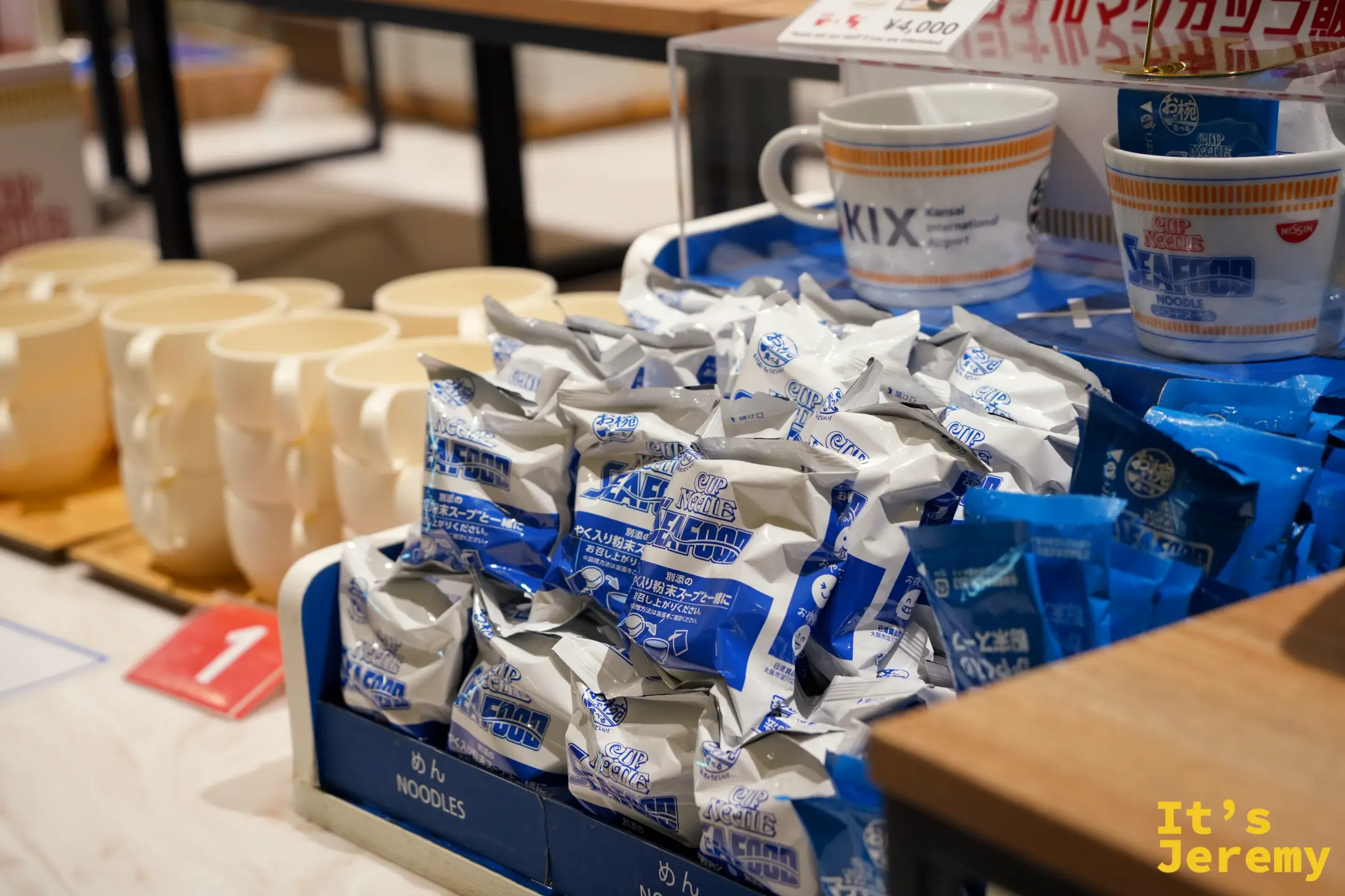 藍色包裝的海鮮口味麵體與湯包，後方展示 KIX 原創馬克杯